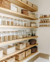 A clean boho pantry with light oak open shelving, white walls, and a variety of clear glass jars with bamboo lids. Woven rattan baskets and light wooden crates neatly store pantry items, while a subtle off-white runner adds texture to the floor. — styling and décor detail