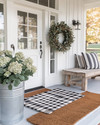 A charming farmhouse exterior with a white front door, layered buffalo check and coir doormats, and a dried eucalyptus wreath. Galvanized metal planters with hydrangeas flank the entrance, beside a distressed wooden bench with a striped throw pillow. — close-up of textures and materials