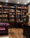A traditional library featuring dark walnut built-in bookshelves filled with books, a deep oxblood leather Chesterfield sofa, and a classic banker's lamp on a mahogany desk. Warm ambient lighting from recessed ceiling lights and a bronze chandelier creates a refined glow. — alternate angle