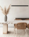 A modern dining room featuring a bleached oak dining table with a live edge, woven rattan dining chairs, and a large, textured cream area rug. A matte black linear pendant light hangs above, and a minimalist ceramic vase with dried pampas grass adorns the table. — close-up of textures and materials