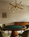 A mid-century modern game room featuring a walnut poker table with a dark green felt top, surrounded by olive velvet low-slung armchairs, illuminated by a brass Sputnik chandelier, with a teak credenza against a soft ivory wall. — styling and décor detail