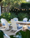 A serene coastal backyard featuring white Adirondack chairs, a weathered teak dining table, and blue and white striped outdoor cushions under strung Edison bulb lights, surrounded by lush green ornamental grasses and potted sago palms. — alternate angle