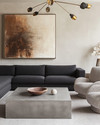Modern great room featuring a low-profile charcoal grey sectional sofa, a large polished concrete coffee table, and a pair of bouclé accent chairs. A dramatic brushed brass chandelier hangs above, complemented by a large abstract painting on a light greige wall. — styling and décor detail