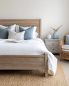 Coastal bedroom with a calming color palette featuring a whitewashed wooden bed frame, crisp white linen bedding, and soft blue throw pillows. A natural jute rug grounds the space, while a rattan armchair and a light grey nightstand with a ceramic vase complete the serene atmosphere. — close-up of textures and materials