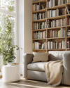 A bright Scandinavian library featuring light oak bookshelves filled with diverse books, a comfortable grey fabric sofa with a beige knit throw, a minimalist white side table, and a large window letting in natural light. — styling and décor detail