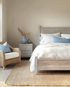 Coastal bedroom with a calming color palette featuring a whitewashed wooden bed frame, crisp white linen bedding, and soft blue throw pillows. A natural jute rug grounds the space, while a rattan armchair and a light grey nightstand with a ceramic vase complete the serene atmosphere. — alternate angle