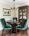 A small traditional dining room featuring a round dark wood pedestal table, upholstered dining chairs in a deep emerald green velvet, and a bronze chandelier hanging above. The walls are painted in a warm cream, and a narrow, glass-front china cabinet stands against one wall, displaying classic dinnerware. — styling and décor detail