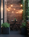 A compact industrial patio featuring a foldable black metal bistro set, string lights with exposed Edison bulbs, and potted green plants in dark grey concrete planters, set against an exposed brick wall. — close-up of textures and materials