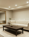 A refined traditional basement living area featuring light greige walls, a tailored neutral linen Chesterfield sofa, and a simple dark wood coffee table. Recessed lighting and a classic brass-finished chandelier illuminate the space, complemented by a large unframed mirror and minimalist decor. — alternate angle