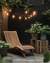 A Scandinavian patio featuring a blend of modern and vintage elements, with a slatted teak lounge chair, a weathered wooden crate side table, and antique ceramic planters filled with green foliage, all under the soft glow of Edison bulb string lights. — styling and décor detail