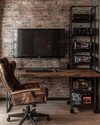 A compact industrial game room featuring a wall-mounted wooden desk with a large monitor, exposed brick wallpaper, and black metal shelving holding retro consoles. A distressed leather gaming chair sits in front, with a small industrial-style bar cart to the side. — close-up of textures and materials