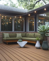 A budget-friendly Mid-Century Modern deck featuring stained pine decking, a low-slung sofa with olive green cushions, and a kidney-shaped side table. Fiberglass planters hold architectural succulents and a large fiddle-leaf fig, all under string lights with exposed bulbs. — alternate angle