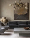 Modern great room featuring a low-profile charcoal grey sectional sofa, a large polished concrete coffee table, and a pair of bouclé accent chairs. A dramatic brushed brass chandelier hangs above, complemented by a large abstract painting on a light greige wall. — alternate angle