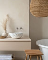 A Japandi bathroom featuring a creamy off-white irregular ceramic vessel sink on a pale oak floating vanity, with a slatted natural teak stool beside a freestanding tub, under a large woven rattan pendant light. — alternate angle