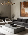 Modern great room featuring a low-profile charcoal grey sectional sofa, a large polished concrete coffee table, and a pair of bouclé accent chairs. A dramatic brushed brass chandelier hangs above, complemented by a large abstract painting on a light greige wall.