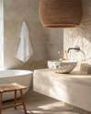 A Japandi bathroom featuring a creamy off-white irregular ceramic vessel sink on a pale oak floating vanity, with a slatted natural teak stool beside a freestanding tub, under a large woven rattan pendant light. — styling and décor detail