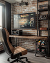 A compact industrial game room featuring a wall-mounted wooden desk with a large monitor, exposed brick wallpaper, and black metal shelving holding retro consoles. A distressed leather gaming chair sits in front, with a small industrial-style bar cart to the side. — alternate angle