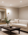 A refined traditional basement living area featuring light greige walls, a tailored neutral linen Chesterfield sofa, and a simple dark wood coffee table. Recessed lighting and a classic brass-finished chandelier illuminate the space, complemented by a large unframed mirror and minimalist decor. — styling and décor detail