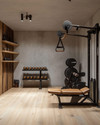 A clean rustic home gym featuring light reclaimed wood flooring, a smooth concrete accent wall, a simple black wrought iron shelf, and minimalist workout equipment with wooden and leather details, bathed in soft ambient lighting. — alternate angle
