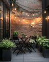 A compact industrial patio featuring a foldable black metal bistro set, string lights with exposed Edison bulbs, and potted green plants in dark grey concrete planters, set against an exposed brick wall. — alternate angle