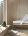 A compact Scandinavian poolhouse interior featuring light birch wood furniture, a white linen throw on a simple bench, and a small round jute rug. A ceramic planter with a green succulent sits on a minimalist shelf, reflecting natural light from a large window. — alternate angle