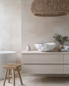 A Japandi bathroom featuring a creamy off-white irregular ceramic vessel sink on a pale oak floating vanity, with a slatted natural teak stool beside a freestanding tub, under a large woven rattan pendant light.