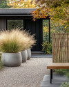 A Scandinavian front yard featuring a pea gravel path, large concrete planters with ornamental grasses, a low woven willow fence, and a slatted wooden bench by the matte black front door. — styling and décor detail