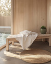 A compact Scandinavian poolhouse interior featuring light birch wood furniture, a white linen throw on a simple bench, and a small round jute rug. A ceramic planter with a green succulent sits on a minimalist shelf, reflecting natural light from a large window.
