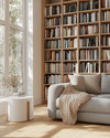A bright Scandinavian library featuring light oak bookshelves filled with diverse books, a comfortable grey fabric sofa with a beige knit throw, a minimalist white side table, and a large window letting in natural light. — alternate angle