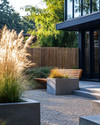 A Scandinavian front yard featuring a pea gravel path, large concrete planters with ornamental grasses, a low woven willow fence, and a slatted wooden bench by the matte black front door.