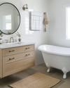 A vintage modern coastal bathroom featuring a white clawfoot tub, a light oak floating vanity, and a round frameless mirror, accented with polished nickel fixtures and a jute bath mat. — styling and décor detail
