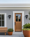 A serene Scandinavian exterior with a light gray facade, crisp white trim, and a natural wood front door. Simple black sconces flank the door, and a minimalist wooden bench sits beside terracotta planters filled with green foliage, all under a bright, clear sky. — alternate angle