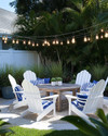 A serene coastal backyard featuring white Adirondack chairs, a weathered teak dining table, and blue and white striped outdoor cushions under strung Edison bulb lights, surrounded by lush green ornamental grasses and potted sago palms. — styling and décor detail