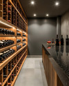 Modern wine cellar with warm grey walls, rift-sawn oak wine racks, and a honed black granite tasting counter. Integrated LED strip lighting illuminates the wine bottles, while recessed spotlights provide ambient glow. — alternate angle