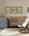 A modern baby and kids room featuring a natural maple crib, a mid-century modern rocking chair with grey linen upholstery, and a vintage wooden toy chest in faded blue. The walls are painted in a warm off-white, complemented by a subtle striped rug and a collection of vintage framed botanical prints.