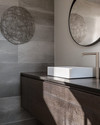 A modern powder room features a floating dark wood vanity with an integrated white quartz sink and brushed nickel faucet, set against large format charcoal grey porcelain tiles. A simple round mirror and abstract metal wall art complete the sleek, resilient design. — styling and décor detail