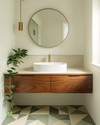 A Mid-Century Modern bathroom featuring a walnut floating vanity with an integrated white sink, brushed brass fixtures, and a large round mirror. The walls are painted a soft off-white, complemented by geometric floor tiles in muted green and cream. A trailing Pothos plant adds a touch of organic warmth. — styling and décor detail