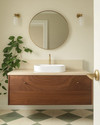 A Mid-Century Modern bathroom featuring a walnut floating vanity with an integrated white sink, brushed brass fixtures, and a large round mirror. The walls are painted a soft off-white, complemented by geometric floor tiles in muted green and cream. A trailing Pothos plant adds a touch of organic warmth. — alternate angle