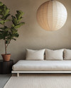 A serene Japandi poolhouse interior featuring a low-slung white oak daybed with natural linen cushions, a dark wenge wood side table, and a large, round paper lantern pendant light. The walls are a soft greige, complemented by a subtle textured rug in cream and a tall, slender fiddle leaf fig plant in a terracotta pot.