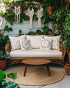 A cozy boho patio featuring a low-profile rattan loveseat with cream cushions, layered with macrame and embroidered pillows. A round jute rug sits beneath a small wooden coffee table, surrounded by various potted plants in terracotta and ceramic planters, including a trailing Pothos in a macrame hanger. — styling and décor detail