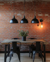 Industrial breakfast nook featuring a reclaimed wood table, black metal chairs, and three matte black dome pendant lights hanging low. An exposed red brick wall provides a textured backdrop, and an exposed Edison bulb sconce adds warm, ambient lighting. — alternate angle