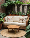 A cozy boho patio featuring a low-profile rattan loveseat with cream cushions, layered with macrame and embroidered pillows. A round jute rug sits beneath a small wooden coffee table, surrounded by various potted plants in terracotta and ceramic planters, including a trailing Pothos in a macrame hanger. — alternate angle