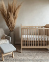 A serene Japandi baby room with a light wood crib, a soft grey glider, and a neutral rug, accented by a tall dried pampas grass arrangement in a ceramic vase and a subtly patterned throw blanket. — alternate angle