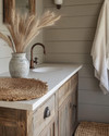 A cozy rustic powder room featuring a reclaimed wood vanity with an off-white quartz countertop, an oil-rubbed bronze faucet, and shiplap walls painted in a soft greige. A woven jute rug sits on the floor, and a ceramic vase with dried pampas grass adorns the counter. — alternate angle