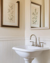 A traditional powder room featuring a white pedestal sink, polished nickel faucet, and beadboard wainscoting painted in a warm off-white. A large framed mirror hangs above, reflecting a botanical print on the opposite wall, all under soft, warm lighting. — styling and décor detail