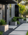 A modern exterior featuring a fresh coat of deep charcoal grey paint on the siding, a contrasting matte black front door, and sleek brushed nickel house numbers. The pathway combines light grey concrete pavers with dark pea gravel, and large concrete planters flank the entrance with sculptural green plants.