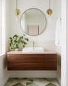 A Mid-Century Modern bathroom featuring a walnut floating vanity with an integrated white sink, brushed brass fixtures, and a large round mirror. The walls are painted a soft off-white, complemented by geometric floor tiles in muted green and cream. A trailing Pothos plant adds a touch of organic warmth. — close-up of textures and materials