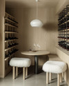 A small Scandinavian wine cellar with light ash wood shelving, a light gray concrete floor, and matte black metal wine racks. A minimalist white pendant light hangs over a small, round light wood tasting table with two simple stools covered in white sheepskin throws. — close-up of textures and materials