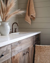 A cozy rustic powder room featuring a reclaimed wood vanity with an off-white quartz countertop, an oil-rubbed bronze faucet, and shiplap walls painted in a soft greige. A woven jute rug sits on the floor, and a ceramic vase with dried pampas grass adorns the counter. — styling and décor detail