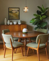 A cozy Mid-Century Modern dining room featuring a round walnut dining table with four upholstered chairs in a muted olive green boucle fabric. A George Nelson bubble lamp hangs above, casting a soft glow. A vintage teak credenza against the wall displays ceramic vases and a large Fiddle Leaf Fig plant. — close-up of textures and materials